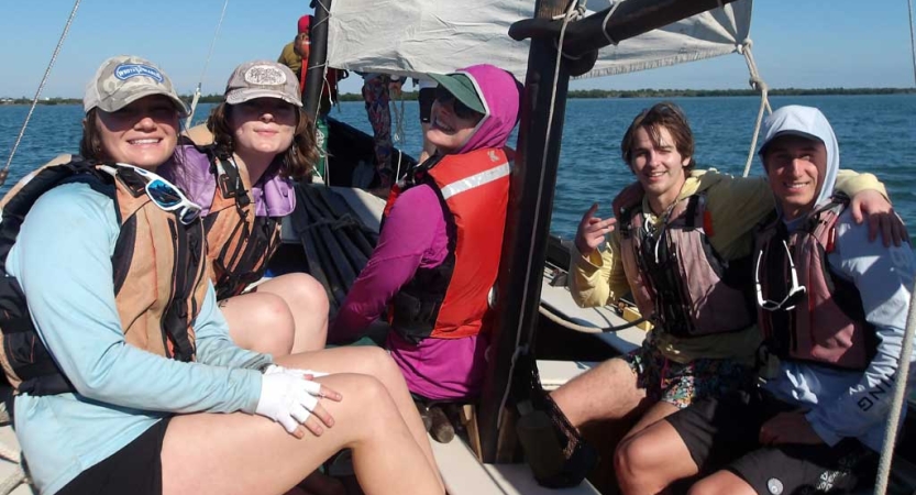 a group of students wearing life jackets sit on a sailboat floating on calm blue water